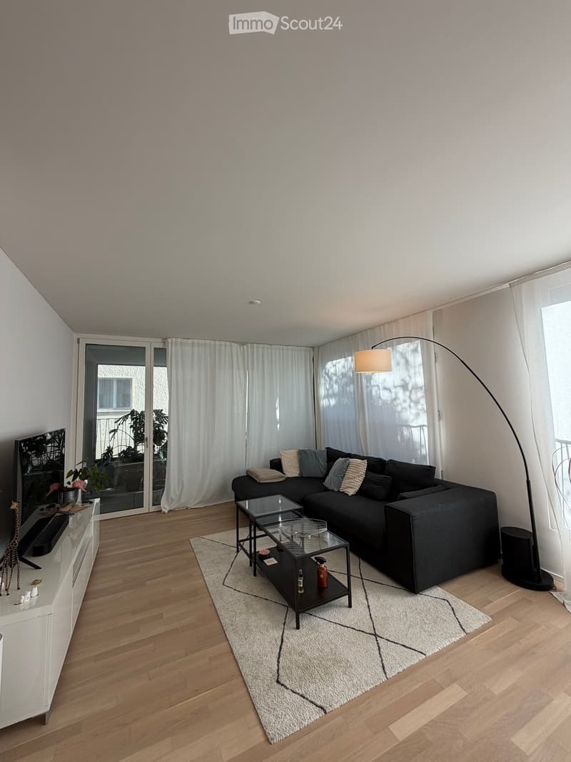 Modern living room with a black sectional sofa, glass coffee table, floor lamp, television on a sideboard, and large windows with white curtains.