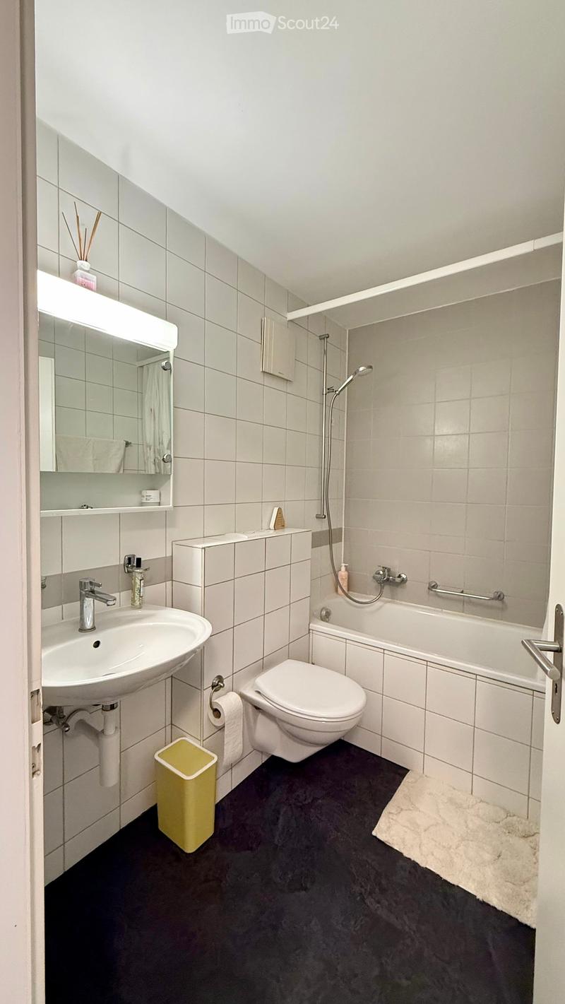 Modern bathroom with white tiles, a sink, toilet, bathtub, showerhead, and a mirror. Includes a yellow trash bin and a white towel on the floor.
