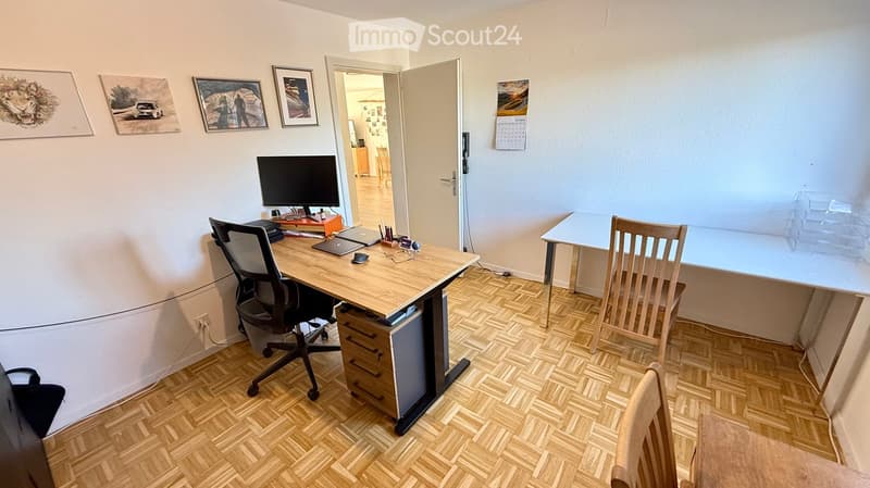 A furnished room with a wooden desk, ergonomic chair, laptop, screen, and a second table with a chair. The room has wooden flooring and several framed pictures on the wall.