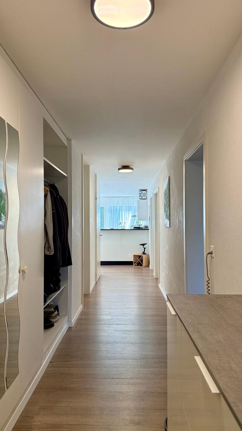 Modern hallway with wooden flooring, white walls, built-in closet with mirrored doors, and recessed lighting. A small table and chair are visible at the end of the hallway.