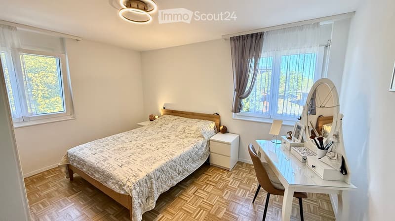Cozy bedroom with a wooden bed, nightstand, vanity desk, and large windows with curtains.