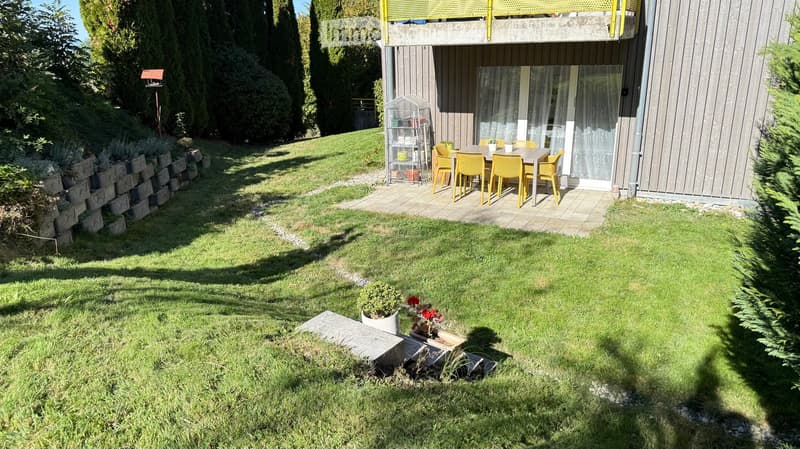 Well-maintained garden with a raised patio area featuring a dining set, surrounded by lush greenery and a modern house with a balcony.