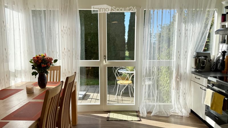 Bright, airy room with large windows and sheer curtains, featuring a dining table with chairs, a vase of flowers, and a modern kitchen area with appliances.
