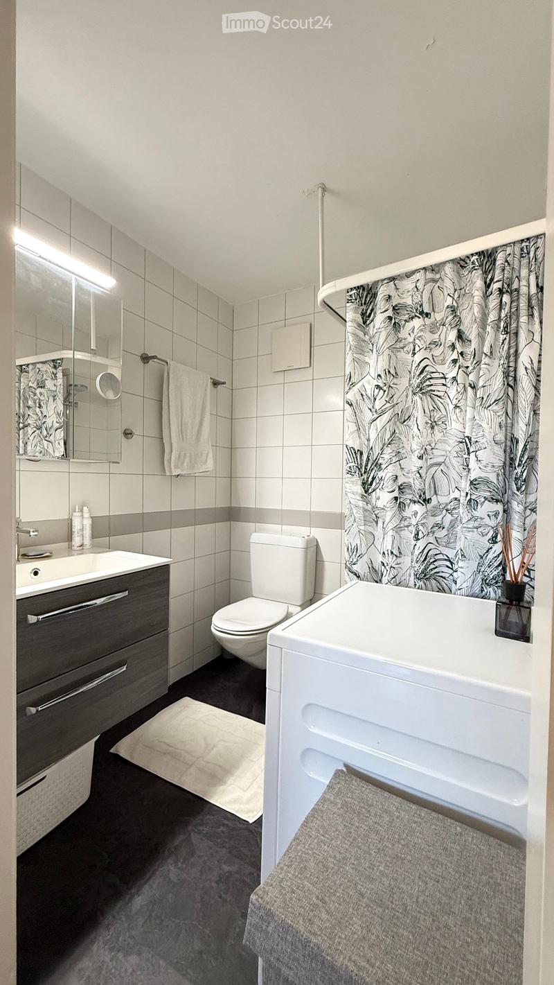 Modern bathroom with white tiles, a dark grey cabinet, a bathtub, a toilet, a sink, and a mirror. Features a floral shower curtain and a towel on the floor.
