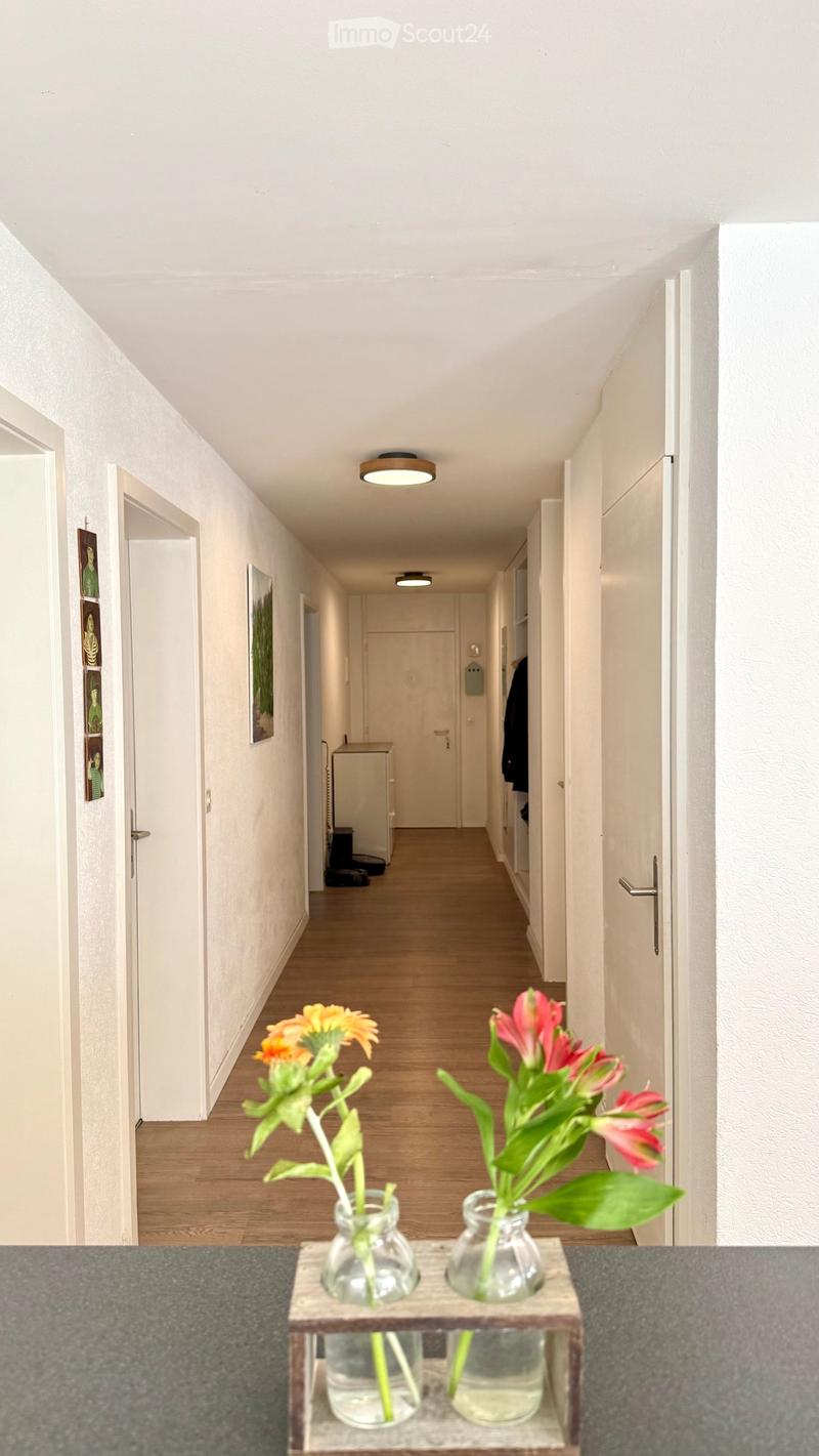 A long, narrow hallway with light wooden flooring, white walls, and recessed lighting. Two vases with flowers are placed on a small wooden stand at the foreground.
