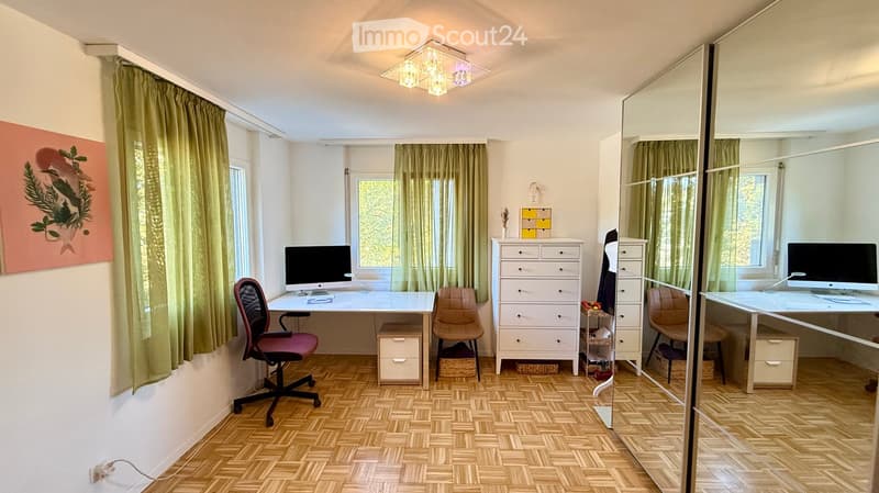 A well-lit room with wooden flooring, a large mirror, and a desk setup with a computer and chair. The room features a drawer unit, a painting on the wall, and green curtains on the windows.