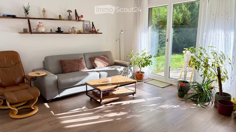 Modern living room with a grey sofa, wooden coffee table, leather armchair, and potted plants near large windows.