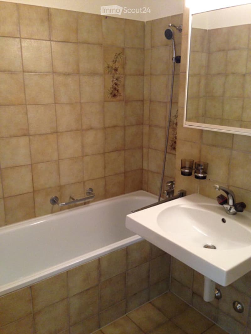 Bathroom with beige tiled walls and floor, a white bathtub, a white sink with a chrome faucet, a mirror above the sink, and a showerhead with a handheld shower attachment.