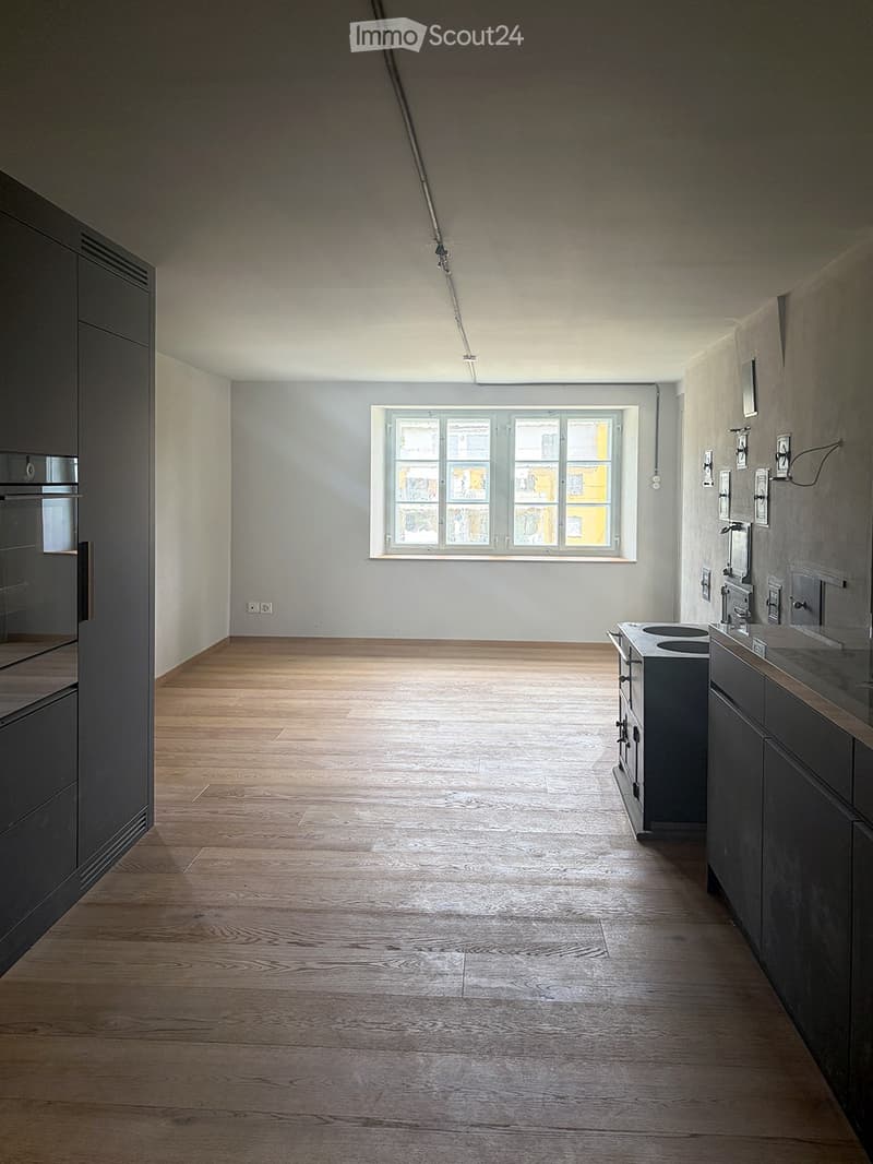 Empty room with wooden flooring, white walls, and a large window letting in natural light. Minimalist design with no furniture or appliances.