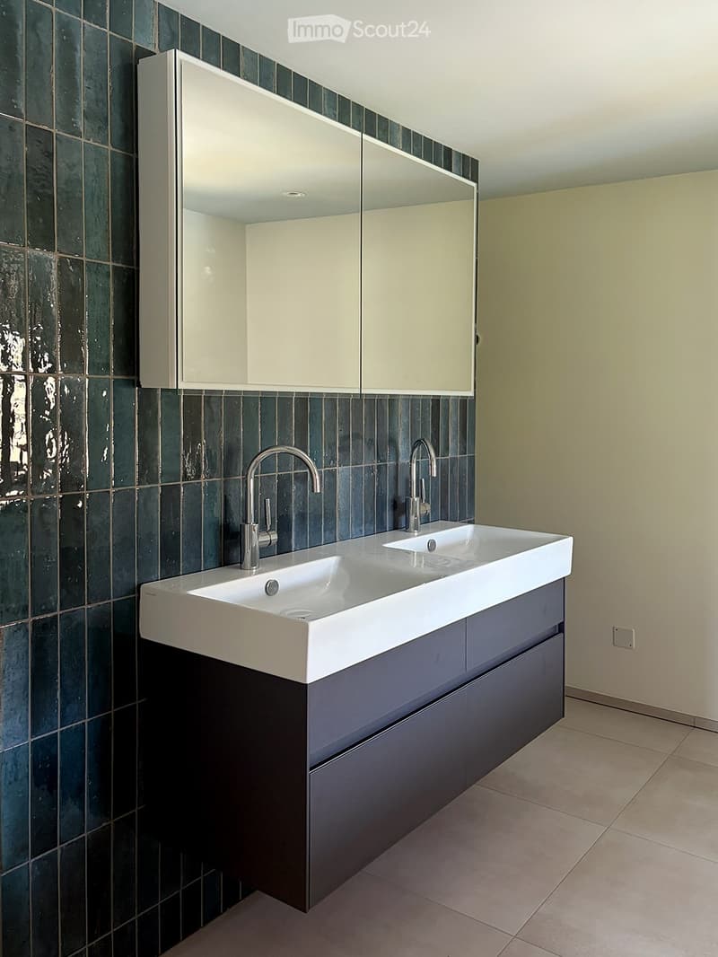 Modern bathroom with a double sink, sleek faucets, a large mirrored cabinet, and dark green tiled walls.