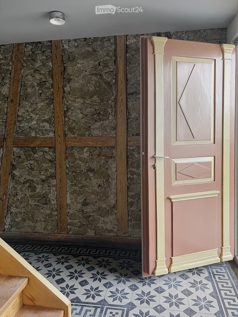 A hallway with a stone wall, wooden beams, and a pink door with decorative molding. The floor features a patterned tile design with geometric shapes and floral motifs.
