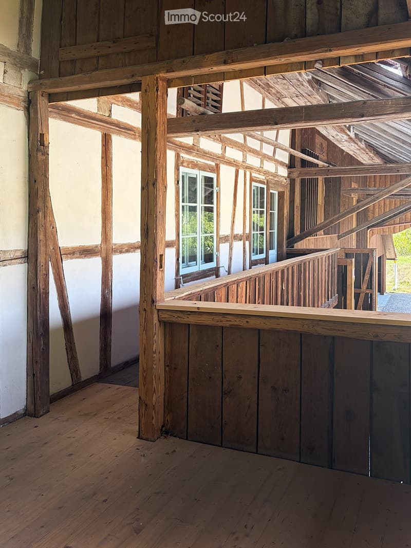 Empty wooden room with exposed beams, wooden floor, and a staircase leading to an upper level. The walls are partially finished with wooden paneling and plaster.