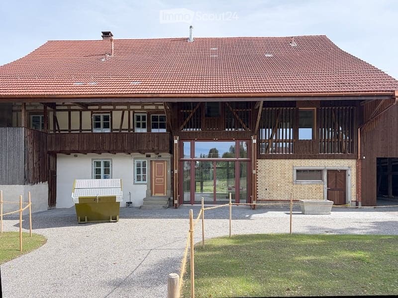 Large two-story building with a red-tiled roof, multiple windows, and a central entrance with a glass door. The structure includes a wooden balcony and a covered porch area.