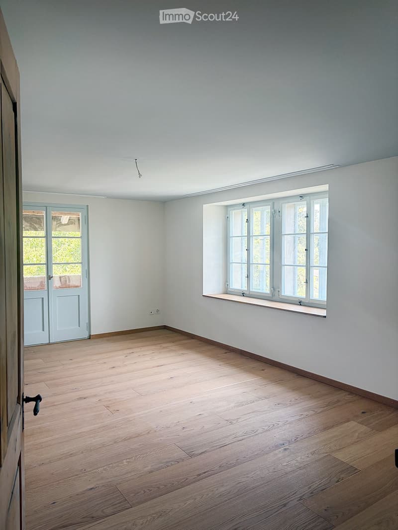 Empty room with light grey walls, wooden flooring, and large windows with white frames. A door with glass panels is visible on the left side.