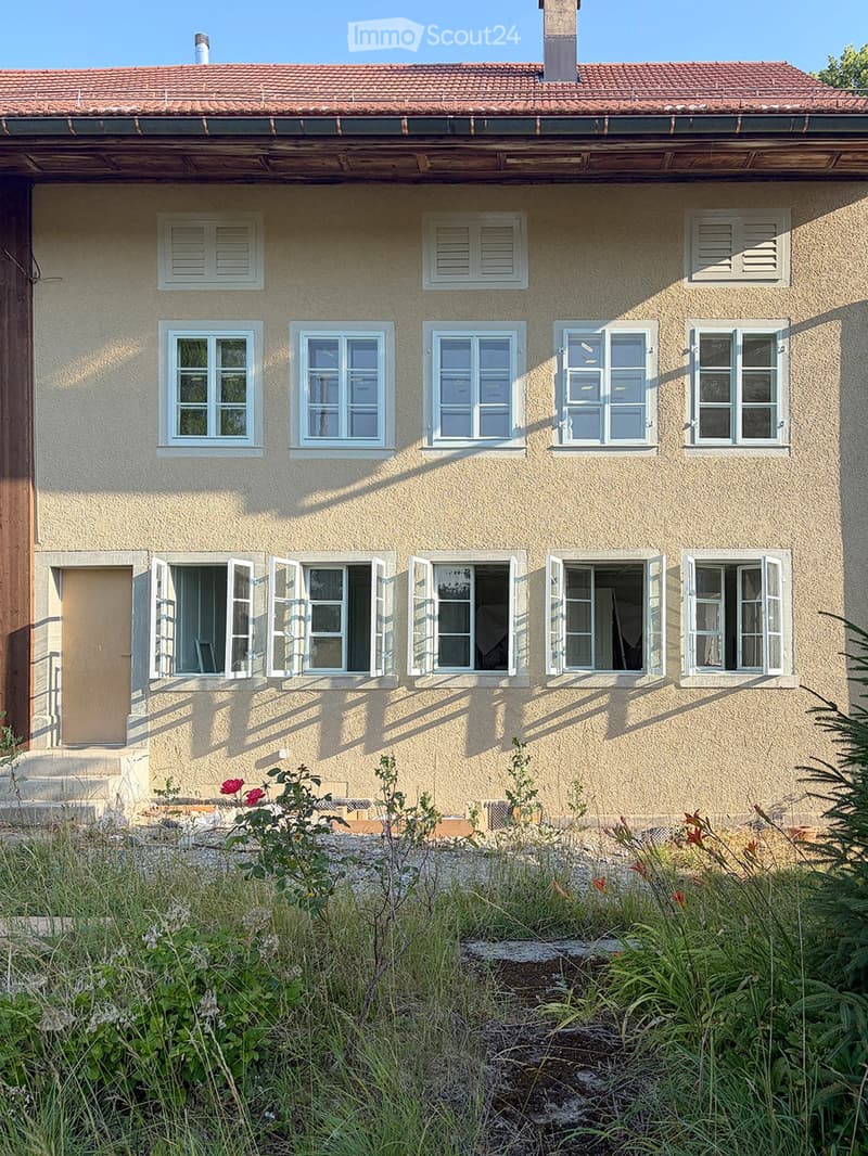 Two-story house with beige exterior, multiple windows, and a tiled roof. The ground floor has visible interior spaces through open windows, and the front yard is overgrown with grass and weeds.