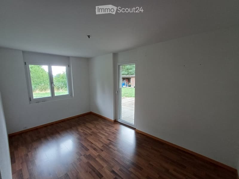 Empty room with wooden flooring, white walls, and a large window with a view of greenery. A sliding glass door leads to an outdoor patio.