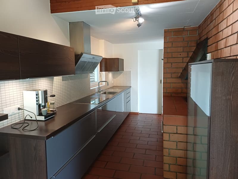 Modern kitchen with dark wooden cabinets, a stainless steel sink, a coffee maker on the counter, and a brick accent wall. The space features a tiled floor and a visible doorway leading to another area.