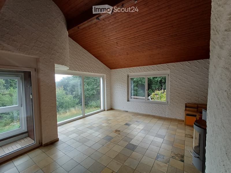 Empty room with tiled floor, exposed wooden ceiling, large windows, and a stone wall. No furniture or equipment present.