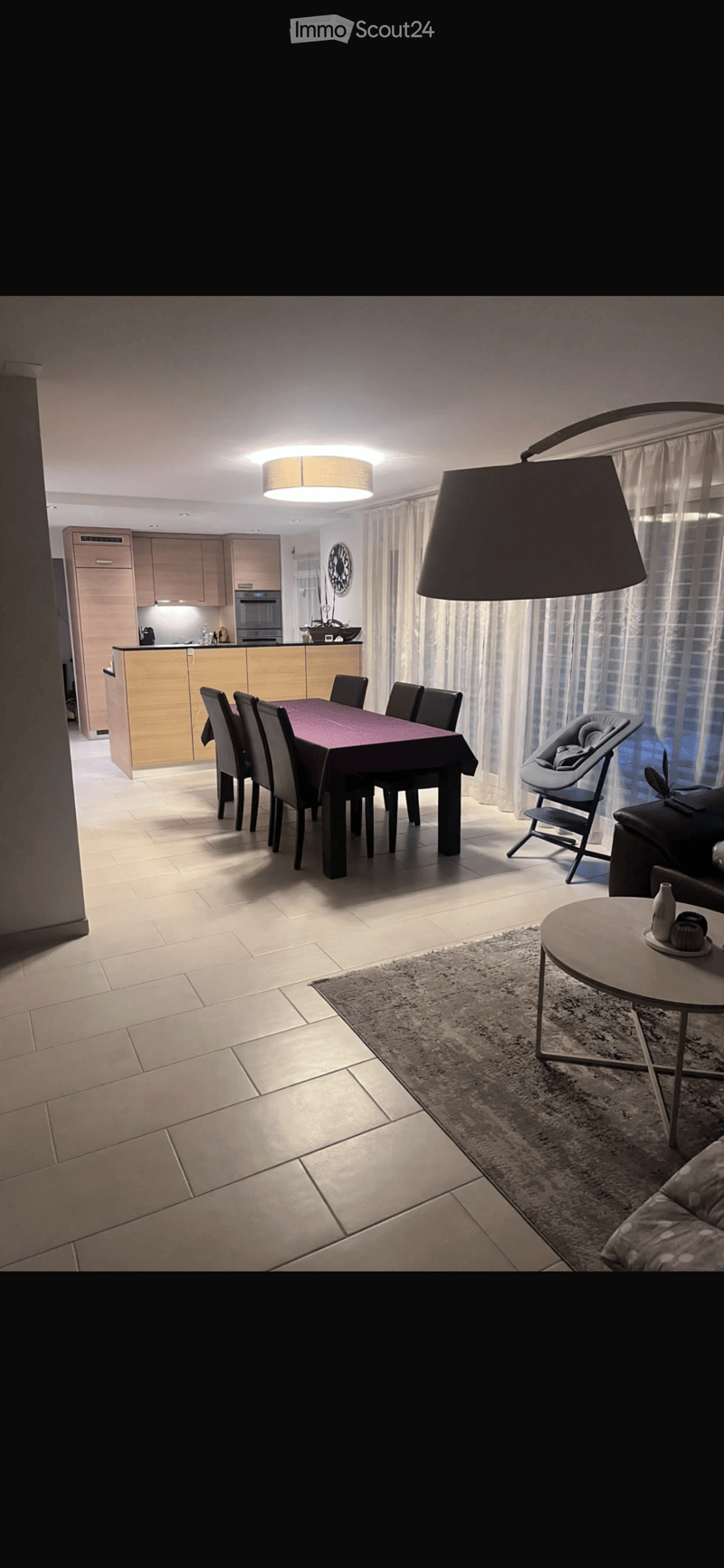 Open-plan living and dining area with a sofa, coffee table, dining table with chairs, baby chair, and a modern kitchen with cabinets, oven, and refrigerator.
