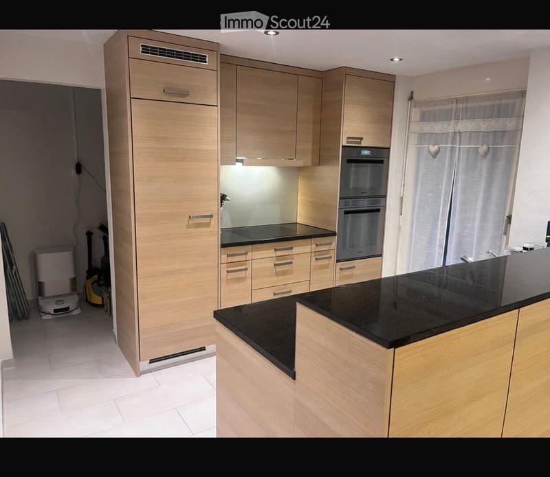 Modern kitchen with light wood cabinets, black countertop, built-in oven, and refrigerator. Features sleek design and ample storage space.
