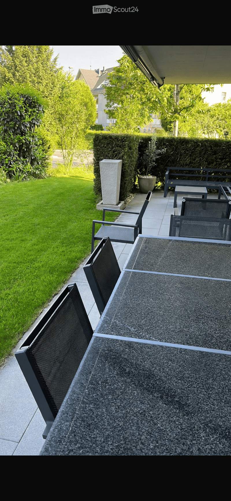 Modern balcony with a stone table, black chairs, and a view of a well-maintained garden with hedges and a lawn.