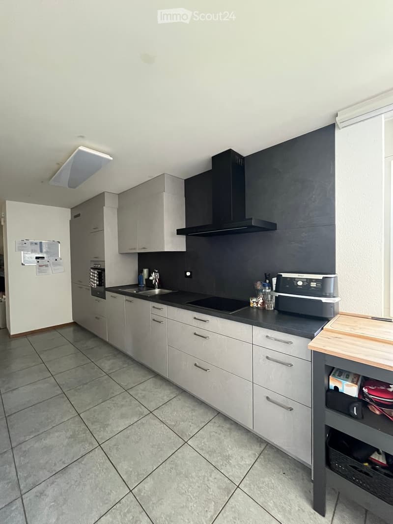 Modern kitchen with grey cabinets, black countertops, and stainless steel appliances including a sink, oven, and range hood. A wooden cutting board and various kitchen items are visible on the counter.