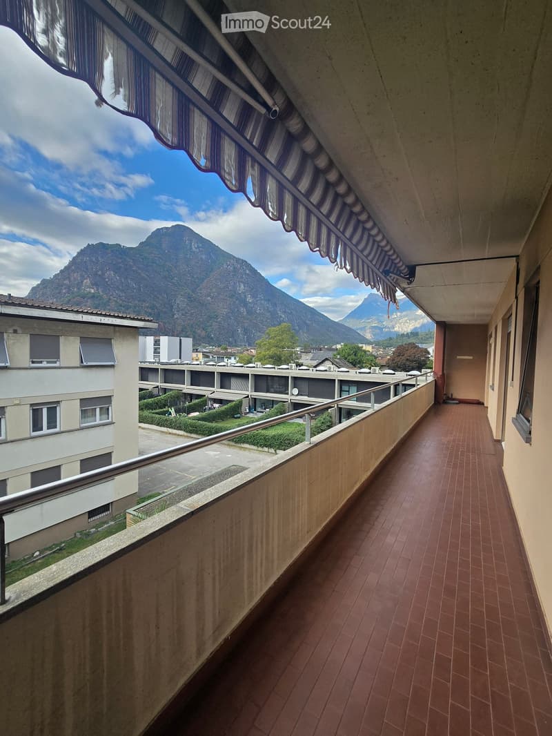 Balcony with red-tiled flooring, metal railing, and a striped awning, overlooking a scenic mountain view and surrounding buildings.
