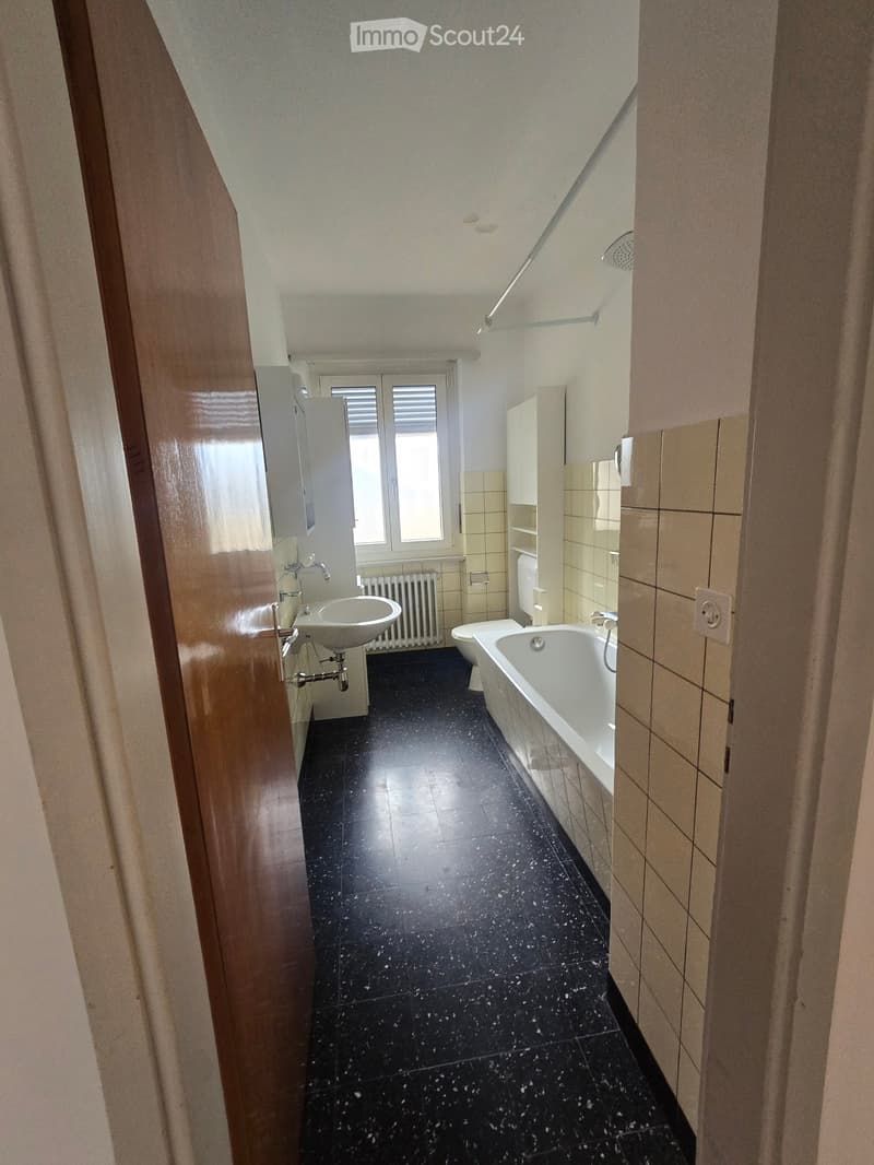 Bathroom with black tiled floor, white bathtub, sink, toilet, and radiator. Light-colored tiles on walls and a window with blinds.