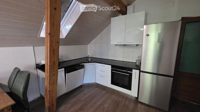 Modern kitchen with white cabinets, stainless steel appliances, black countertops, and a wooden floor. Features include a refrigerator, oven, sink, and a small seating area with a chair.