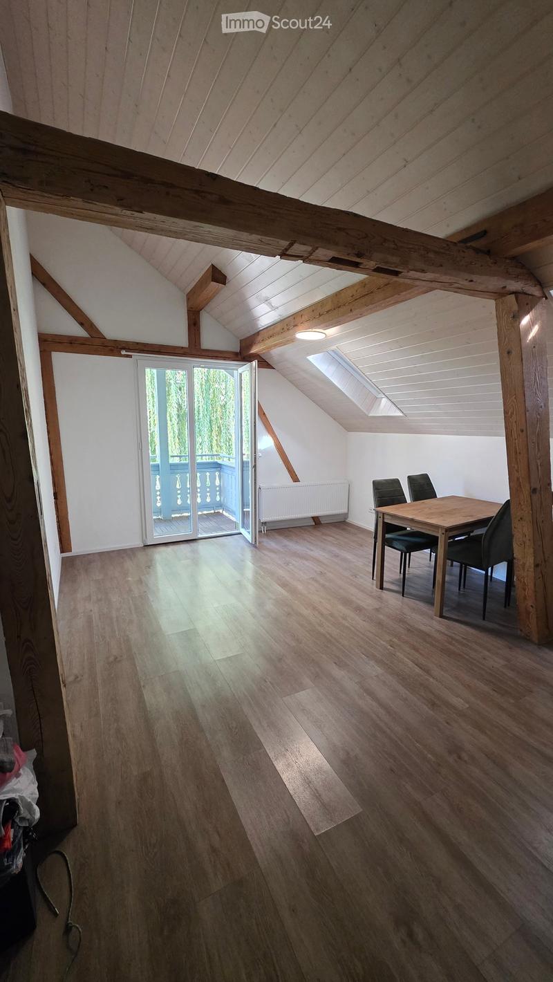 Bright, airy attic space with wooden beams, wooden flooring, and a large skylight. A small dining area with a table and chairs is visible, and a sliding glass door opens to a balcony.