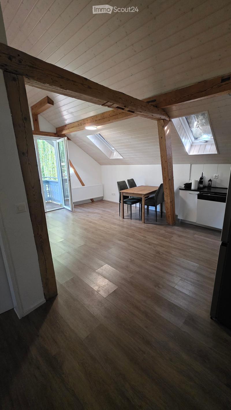 Bright, wooden-floored attic space with exposed wooden beams, skylights, a small dining area with a table and chairs, and a compact kitchen area with a counter and appliances.