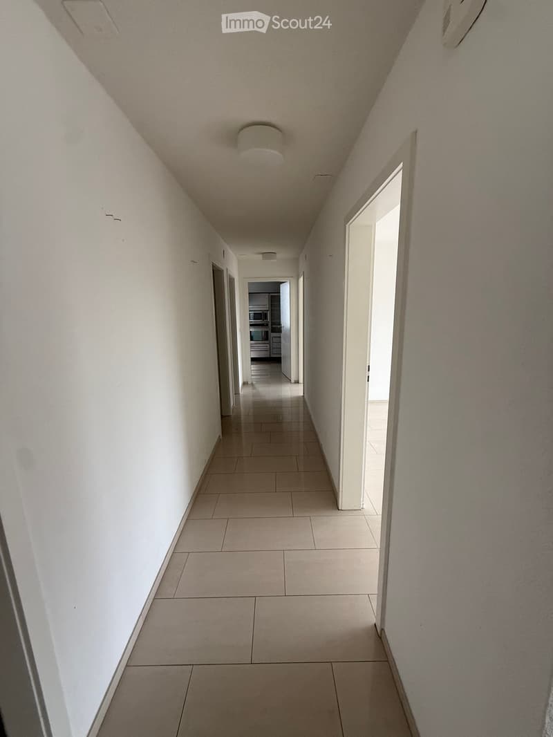 A long, narrow hallway with tiled flooring and white walls, featuring several doors and a visible kitchen area at the end.