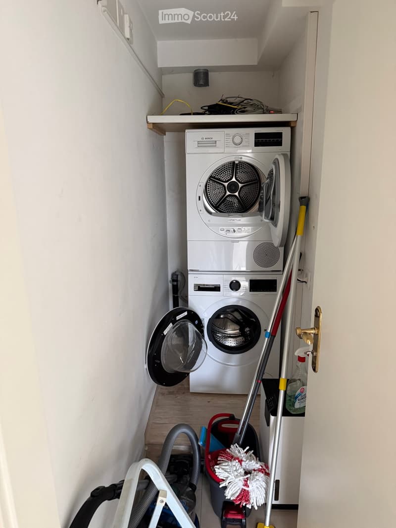 A compact utility room with a stacked washing machine and dishwasher, a mop, vacuum cleaner, and cleaning supplies.