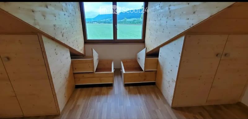 Empty room with wooden flooring, built-in wooden benches along the walls, and a large window offering a scenic view of a field and mountains.