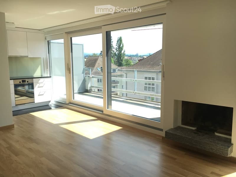 Bright, modern living space with large sliding glass doors opening to a balcony, wooden flooring, fireplace, and a small kitchen area with cabinets and an oven visible.