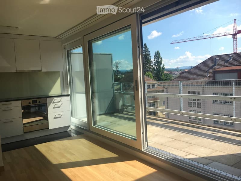 Modern kitchen with light wooden flooring, white cabinets, and an oven. Large sliding glass doors open to a balcony with a view of the city and surrounding buildings.