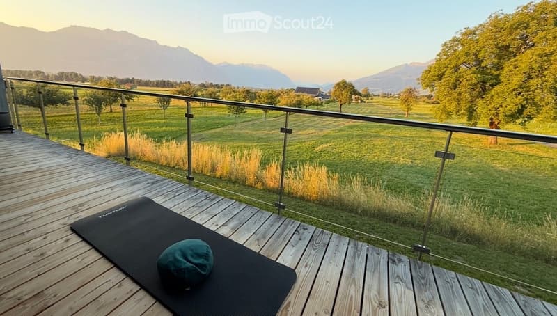 Wooden balcony with glass railing, overlooking a green field and mountains, with a yoga mat and hat placed on the deck.