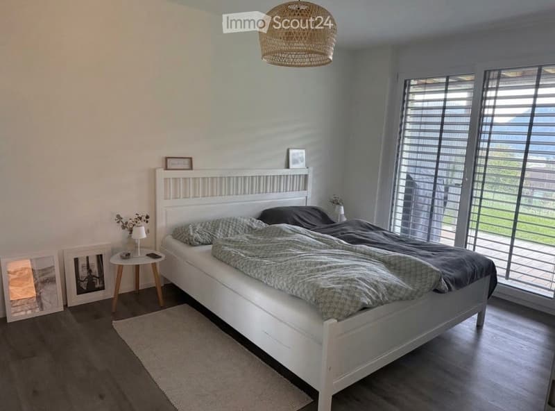 Modern bedroom with a white bed, nightstand, table lamp, and large windows with blinds. The room features a wicker pendant light and a small rug on the floor.