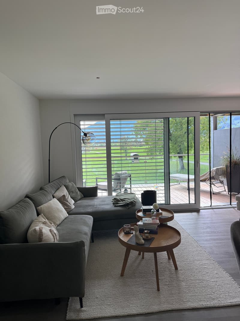 Modern living room with large windows offering a view, a grey sectional sofa, a round wooden coffee table, a floor lamp, and a small table with books and decorative items.