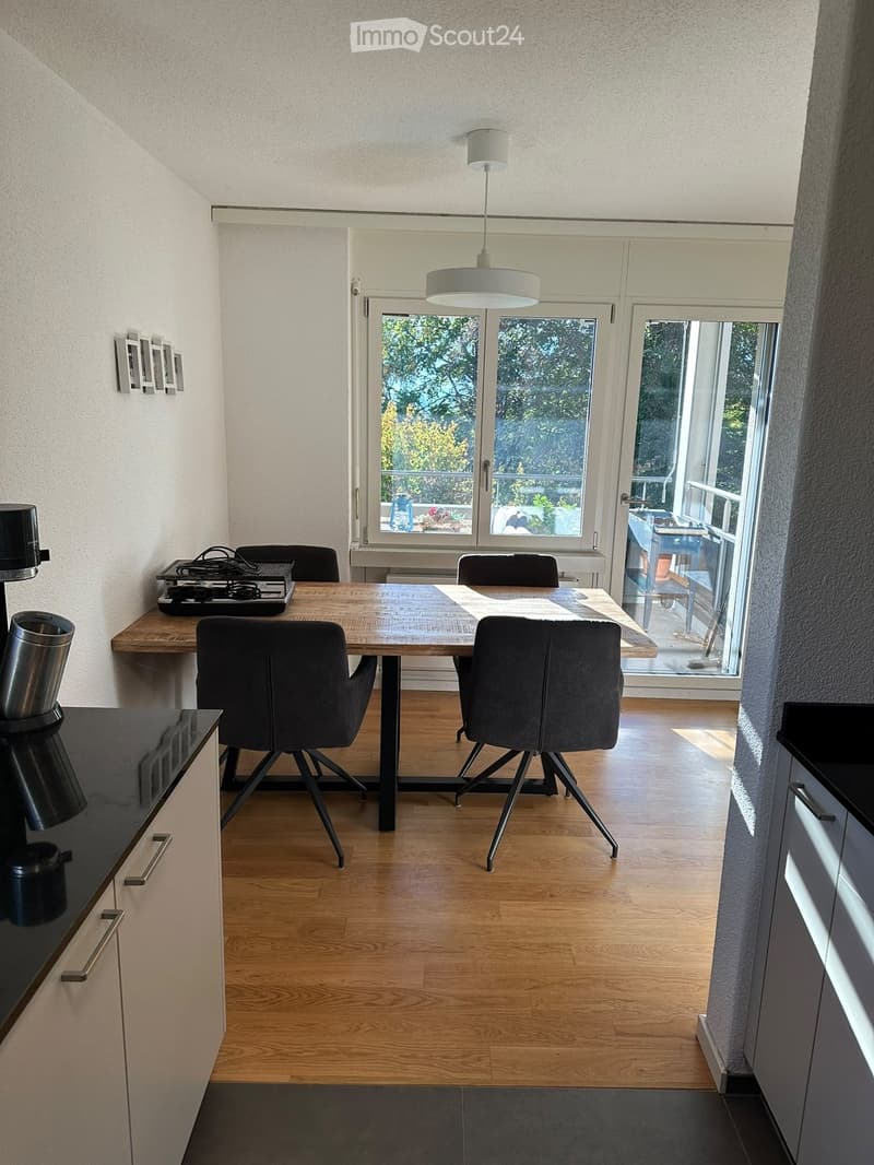 Dining area with wooden table, four chairs, coffee maker on counter, and large windows leading to a balcony.