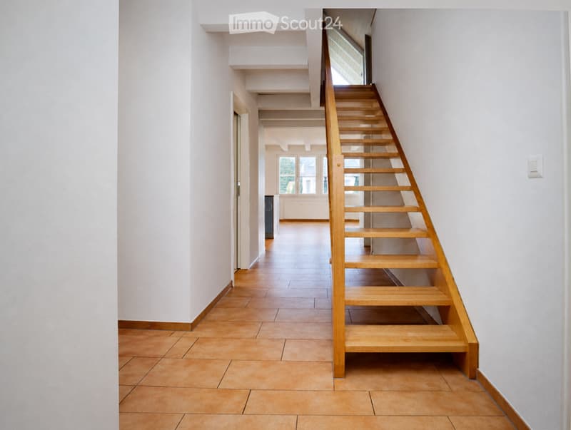 A modern hallway with a wooden staircase leading upwards, tiled flooring, and white walls. The hallway features a door on the left and a window at the end, allowing natural light to enter.