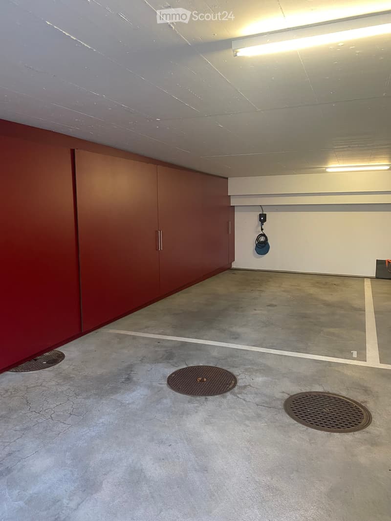 Empty underground parking space with concrete floor, red storage unit, fluorescent lighting, and several manhole covers.