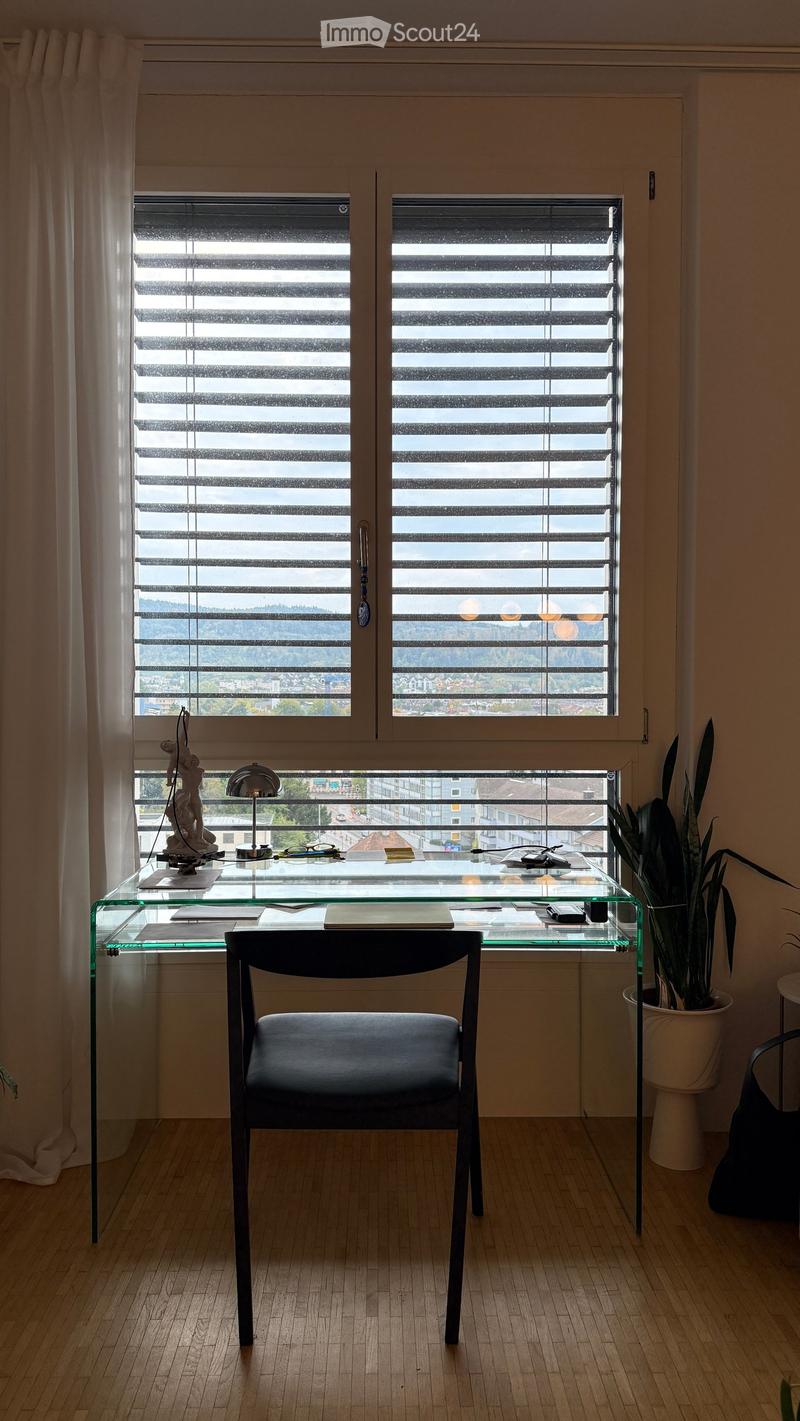 A modern, minimalist room with a glass desk, a black chair, a lamp, a potted plant, and a decorative statue. The room features large windows with blinds and a scenic view of the city.