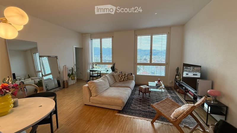 Modern living room with a sectional sofa, coffee table, large mirror, and a view through large windows.