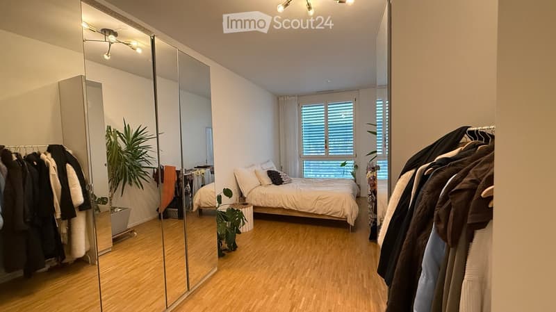 Modern bedroom with a large bed, mirrored wardrobe, and hanging clothes. Features wooden flooring, large window with blinds, and decorative plants.