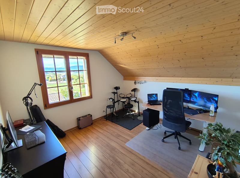 A well-lit attic room with wooden floors and slanted ceilings, featuring two desks with monitors, a drum set, and a guitar, along with chairs and a plant.