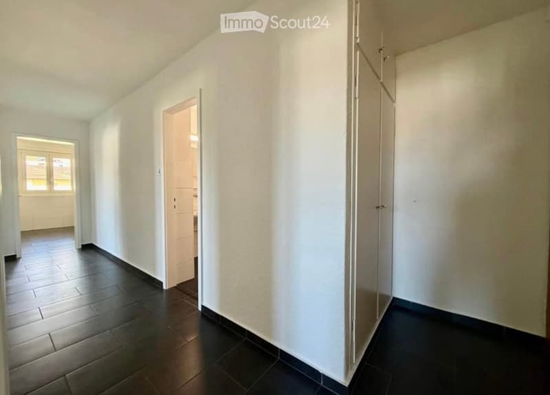 Empty hallway with dark tiled flooring and white walls, featuring an open doorway leading to another room and a window at the far end.