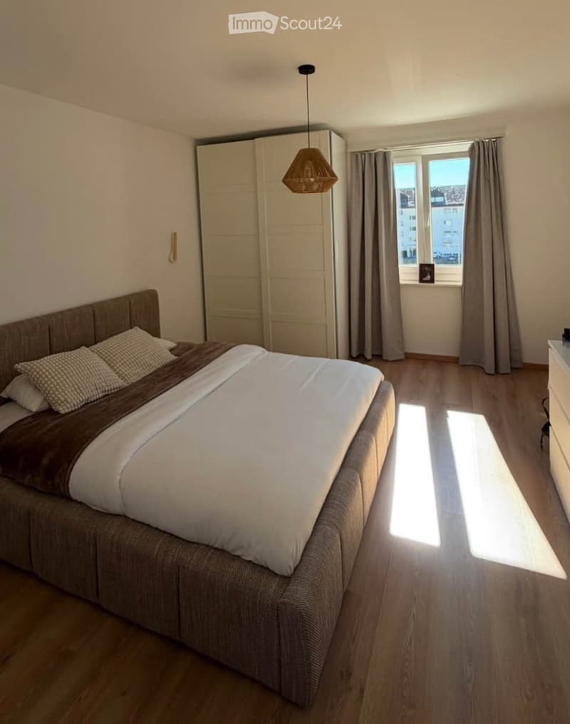 Modern bedroom with a large bed, pillows, and a blanket, a wardrobe, a hanging lamp, and a window with curtains.