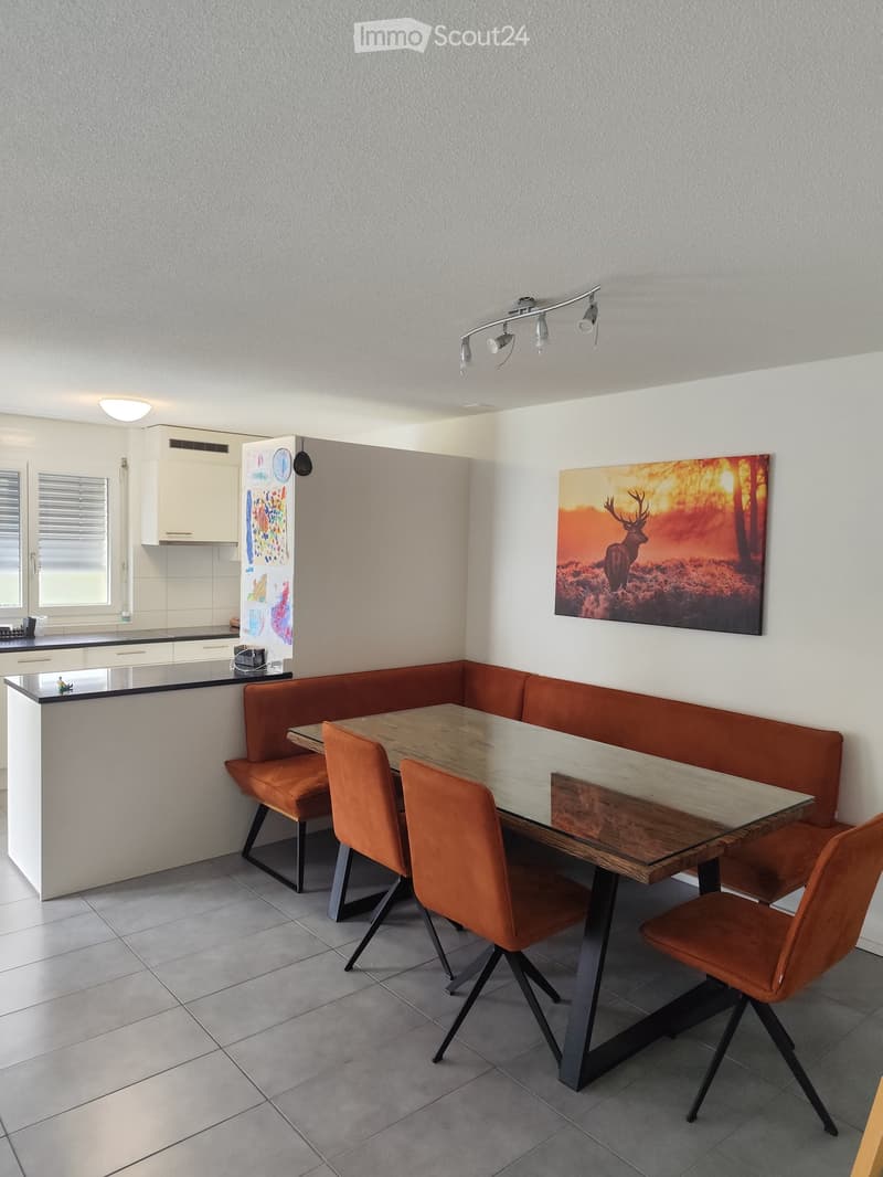 Modern dining area with a wooden table, four orange chairs, and a built-in orange bench seating. The space features a sleek kitchen area with white cabinets and a black countertop, along with a large painting of a deer on the wall.