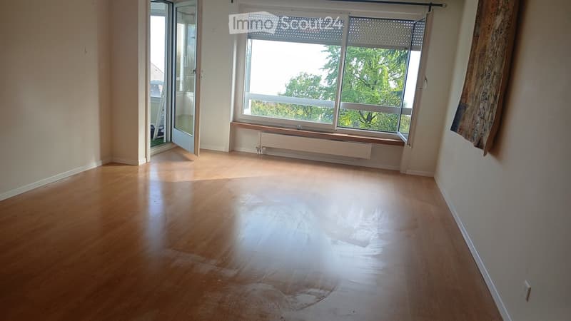Empty room with wooden flooring, large window with blinds, radiator, and an open sliding door leading to a balcony.